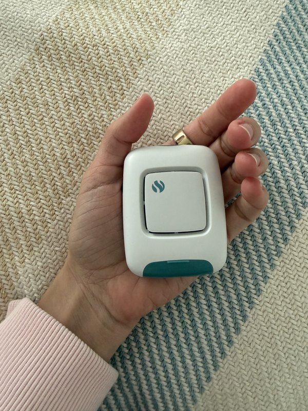 A hand holding a small white FoodMarble AIRE breath tester with a teal mouthpiece, over a woven rug.